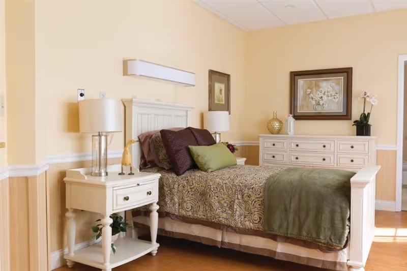 Well-lit bedroom with a made bed, nightstand and dresser, lamps, framed artwork, and soft yellow walls.