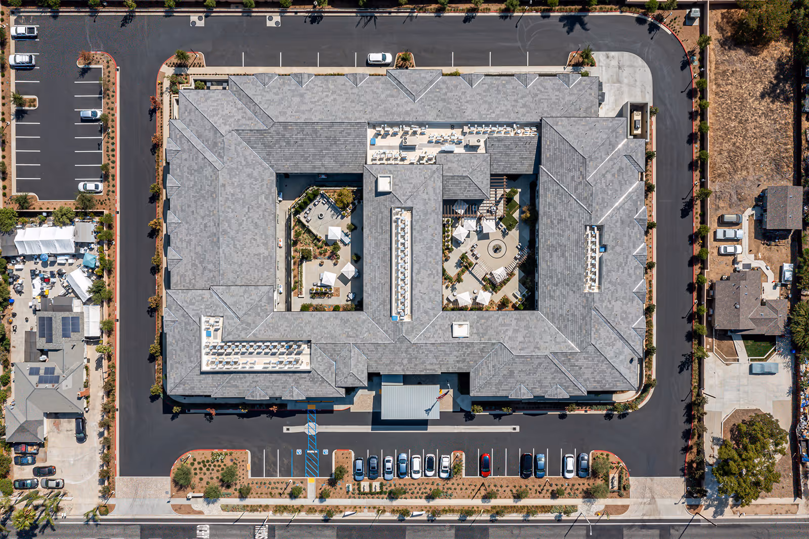 Aerial view of Allara Senior Living facility showing a large rectangular building with a central courtyard featuring outdoor seating areas, umbrellas, and landscaping. Surrounding the building is a parking lot with several parked cars and adjacent residential houses and open land.