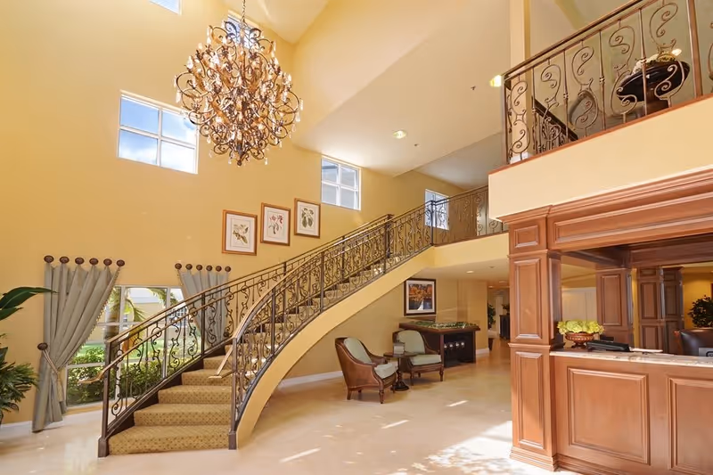 Bright and spacious interior of a senior living facility featuring a curved staircase with ornate wrought iron railing, a large decorative chandelier hanging from the high ceiling, framed botanical artwork on the wall, two armchairs with a small table between them, and a wooden reception desk with a flower arrangement.