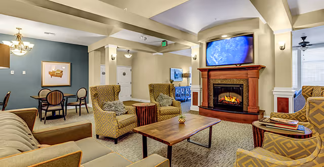 Bright assisted-living common room with sofas and armchairs arranged around a coffee table and a fireplace with a TV above.