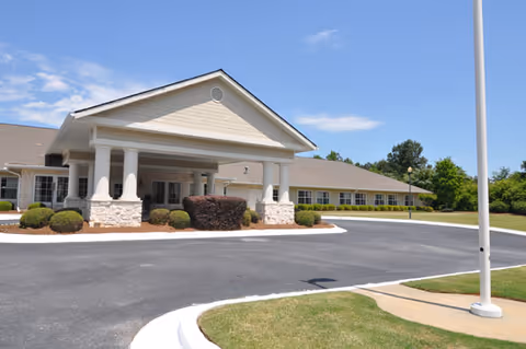 Exterior view of a single-story senior living facility building with a covered entrance supported by white columns, surrounded by landscaped bushes and a paved driveway under a blue sky with some clouds.