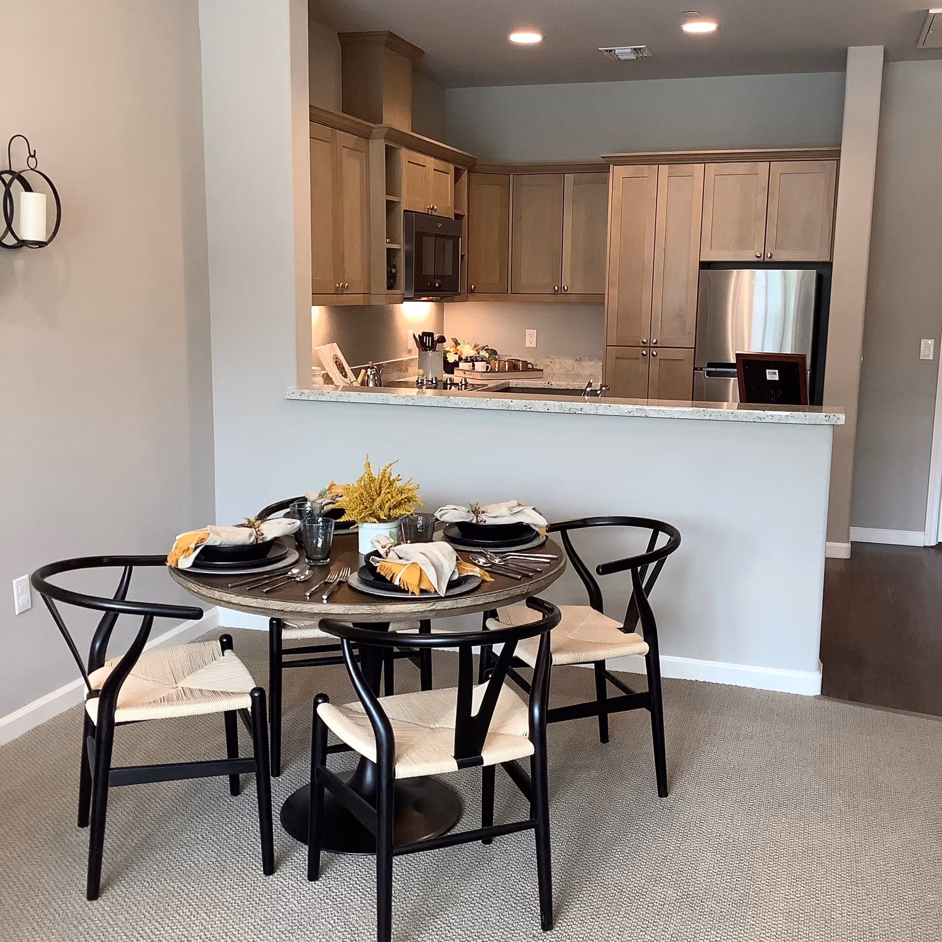 A modern dining area with a round wooden table set for four, featuring black chairs with woven seats. Behind the dining area is a kitchen with light wood cabinets, a stainless steel refrigerator, a microwave, and a granite countertop. The walls are painted light gray and the floor is carpeted in the dining area with hardwood flooring visible in the kitchen.