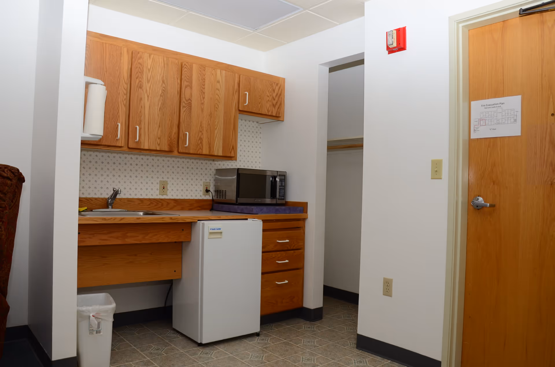 Small kitchenette area with wooden cabinets, a sink, a microwave on the counter, a mini refrigerator below the counter, and a trash can on the floor. There is a wooden door with a fire evacuation plan posted on it and an open closet space next to the kitchenette.