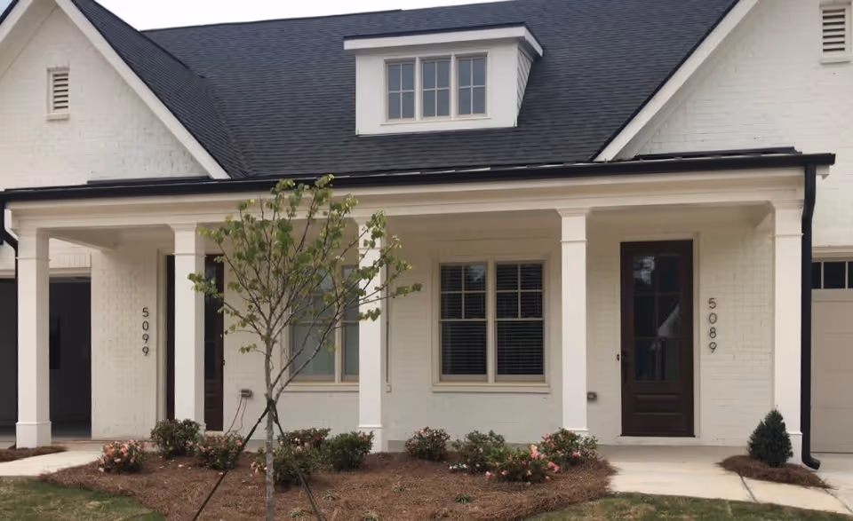 Front exterior view of a white brick building with a black roof, two dark wooden doors numbered 5099 and 5089, a small tree and flower beds in front, and a covered porch supported by white columns.