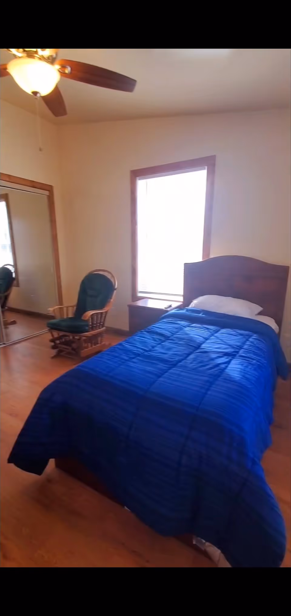 A bedroom with a single bed covered in a blue comforter and a white pillow. Next to the bed is a wooden nightstand under a window with blinds. There is a wooden rocking chair with a green cushion near a mirrored closet door. A ceiling fan with a light fixture is visible on the ceiling.