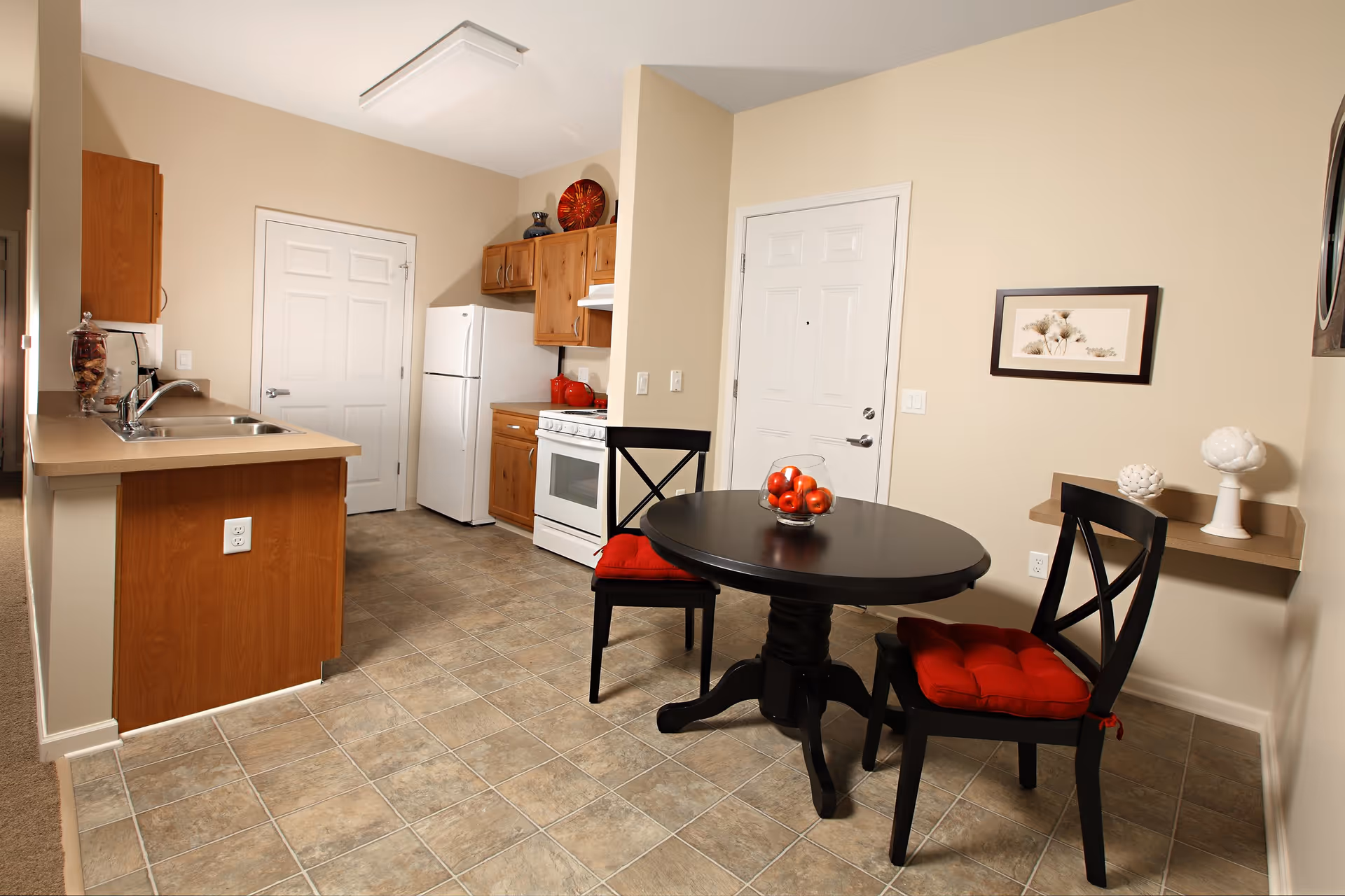A small kitchen and dining area with beige walls and tiled floor. The kitchen has wooden cabinets, a white refrigerator, stove, and a double sink. The dining area features a round black table with two black chairs that have red cushions. A glass bowl with red apples is on the table, and a small shelf on the wall holds decorative items. A framed picture hangs above the shelf.