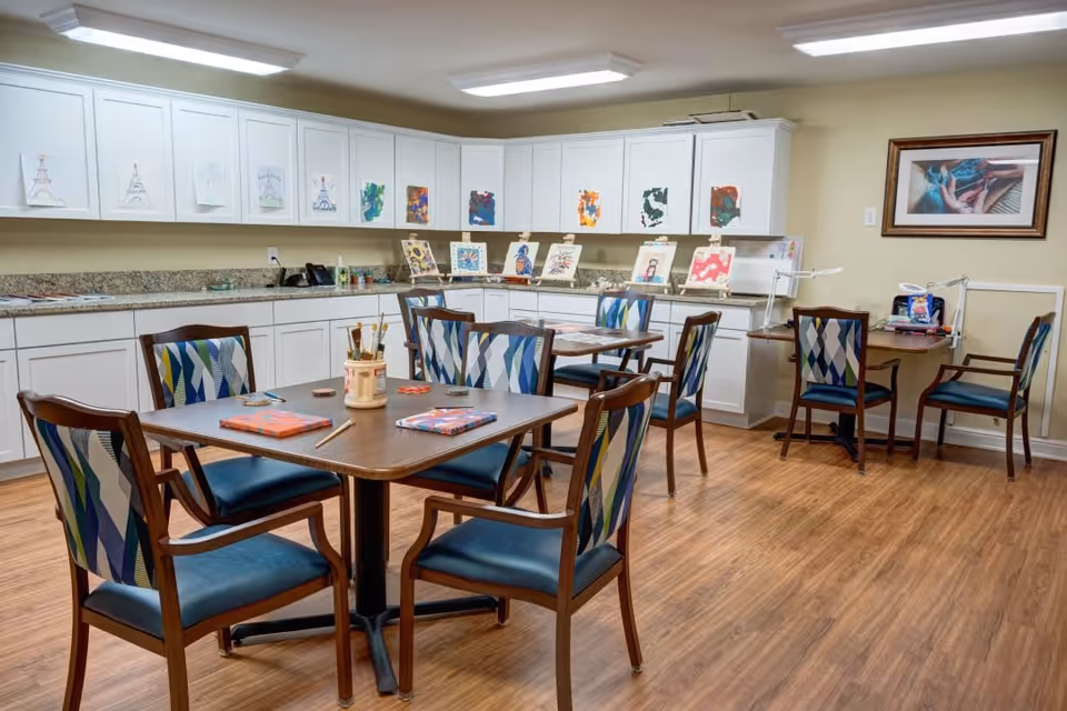 A bright and clean activity room with wooden flooring, several tables and chairs with patterned upholstery, and white cabinets along the back wall. The tables have art supplies like paintbrushes and canvases, and various colorful paintings are displayed on the cabinets and easels. A framed picture hangs on the wall to the right.