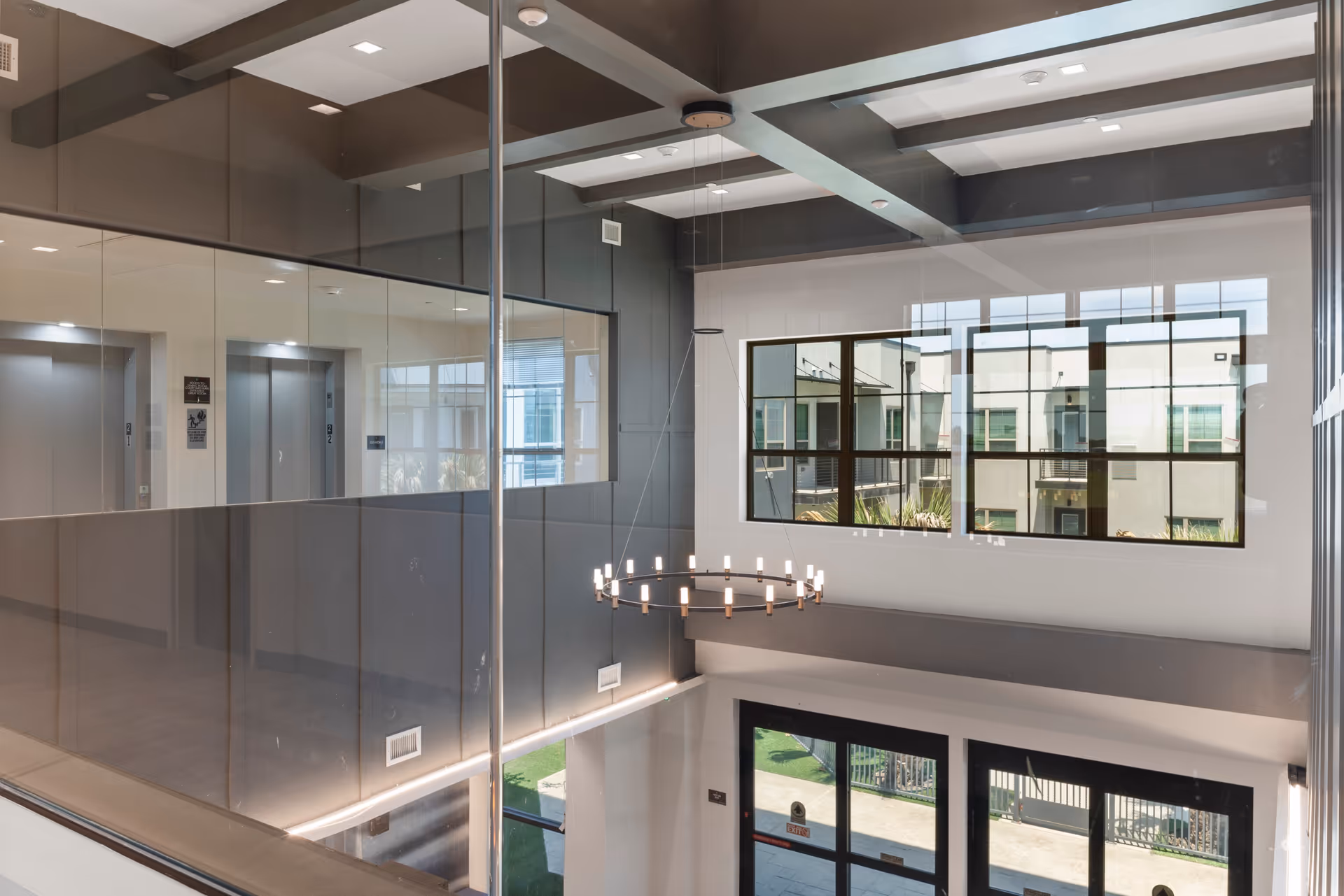 Interior view of a modern senior living facility lobby with large windows, a circular chandelier hanging from the ceiling, glass walls, and an elevator area visible in the background.