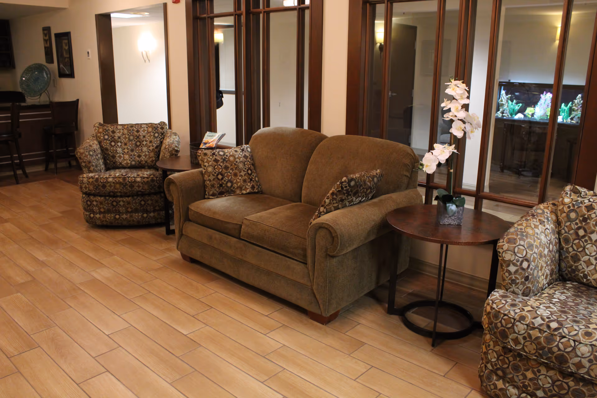 A cozy seating area in a retirement community featuring a brown upholstered loveseat with patterned cushions, two matching patterned armchairs, and two round wooden side tables. One side table holds a white orchid plant. The room has wood-look tile flooring and large windows with wooden frames. In the background, there is a bar area with high chairs and a decorative plate on the counter.