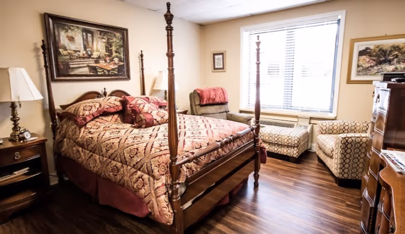 A cozy bedroom with a wooden four-poster bed covered in a patterned bedspread and matching pillows. There is a nightstand with a lamp and clock on the left side of the bed. A cushioned armchair with a red throw blanket is positioned near a window with blinds. Two patterned ottomans and another armchair are near the window. The room has wooden flooring and framed artwork on the walls.