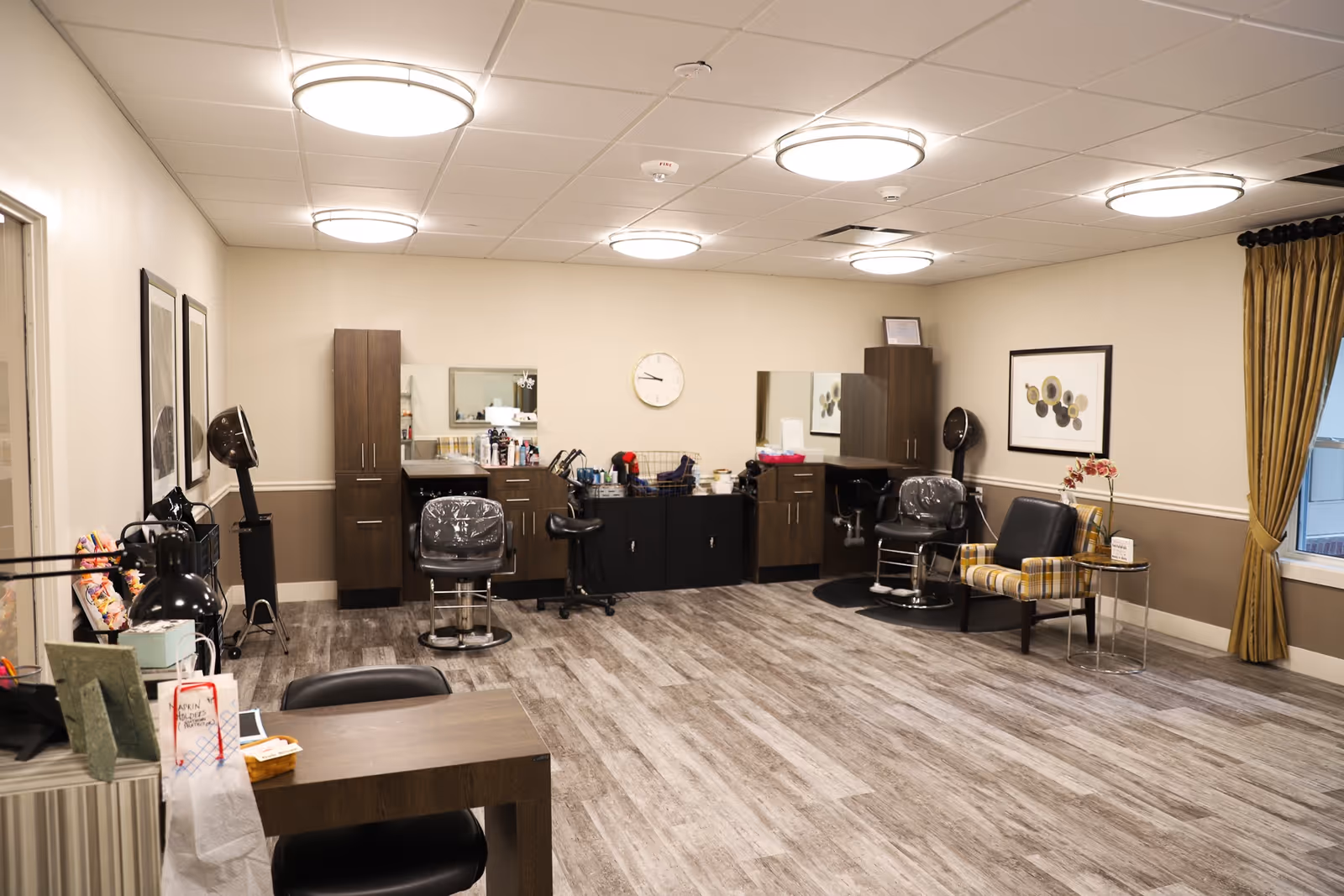 A spacious salon room with two hair styling stations, each equipped with a salon chair, mirrors, and hair dryers. The room has wood-look flooring, beige walls with a darker beige wainscoting, and modern circular ceiling lights. There is a small seating area with a black chair and a plaid armchair next to a window with gold curtains. A clock is mounted on the wall between the two styling stations.
