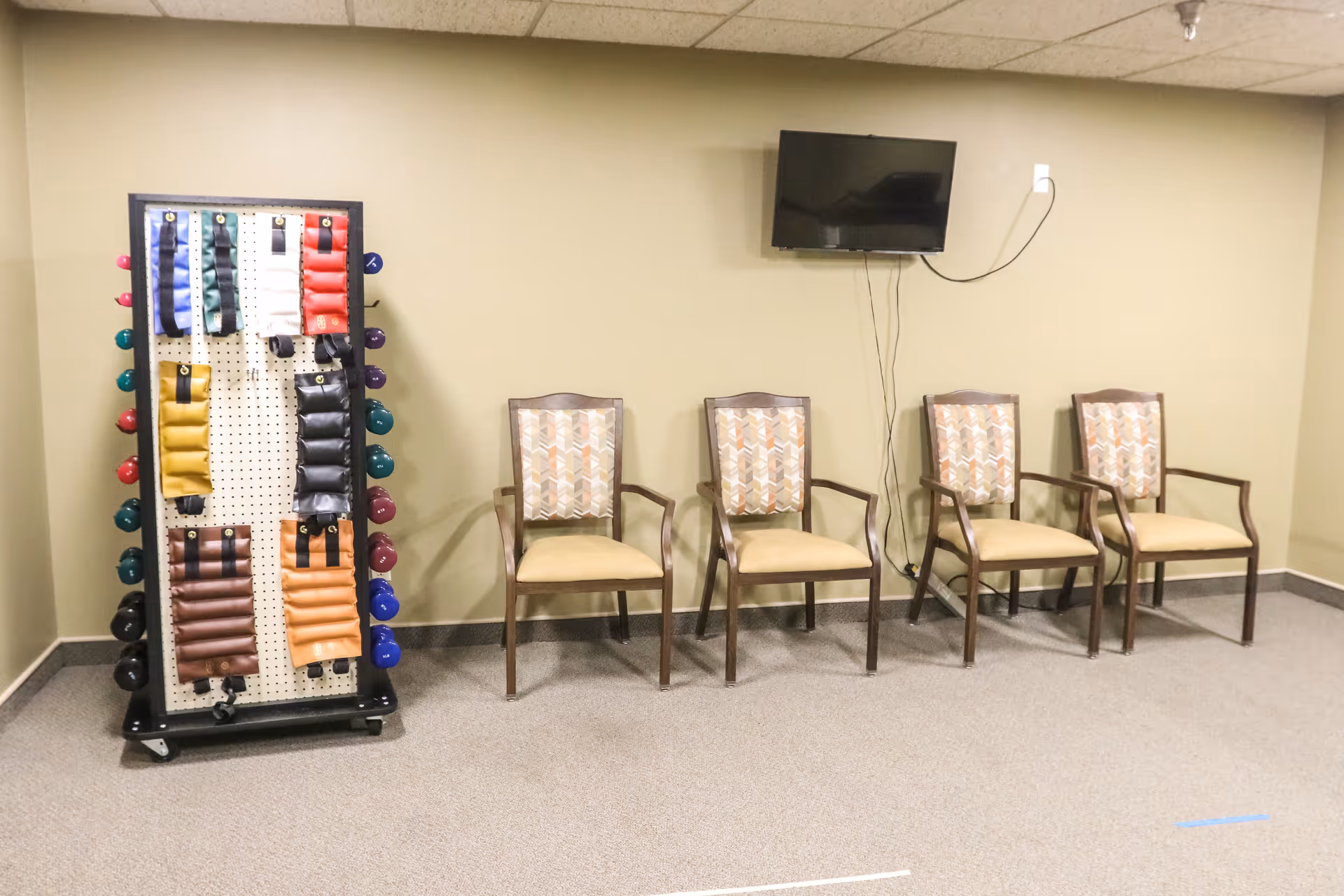 A room with four wooden chairs with patterned cushions arranged in a row against a beige wall. To the left, there is a rack holding various colored exercise weights and resistance bands. A flat-screen TV is mounted on the wall above the chairs with visible cables hanging down.