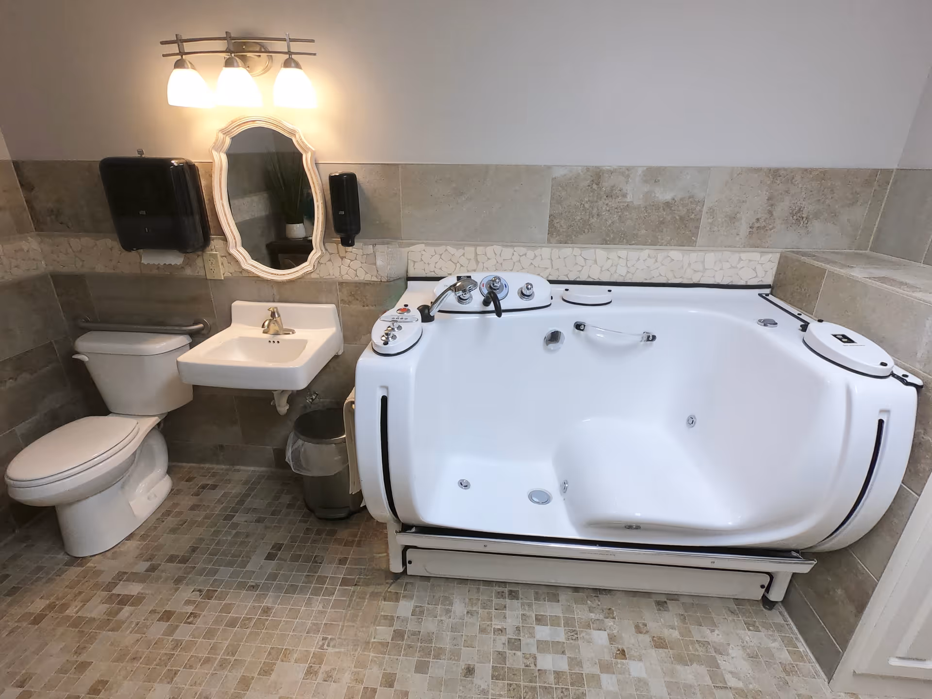 A bathroom with a white toilet, a small white sink with a silver faucet, a decorative mirror above the sink, a wall-mounted soap dispenser, a paper towel dispenser, a trash can, and a large white walk-in bathtub with multiple controls and safety handles. The walls and floor are tiled in neutral tones.