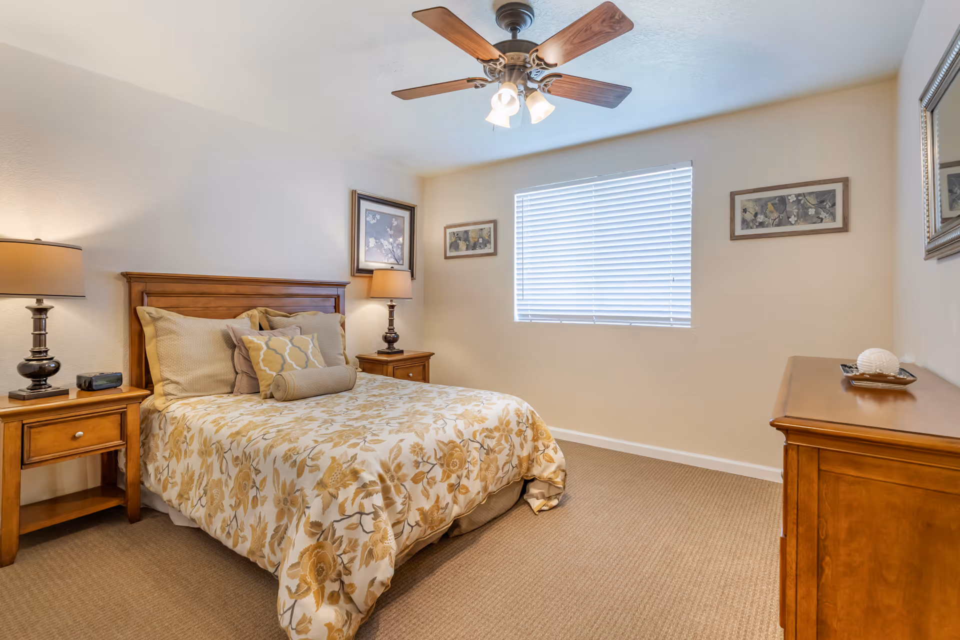 A cozy bedroom with a wooden bed frame and floral patterned bedding in shades of beige and yellow. There are two matching wooden nightstands on either side of the bed, each with a lamp. A ceiling fan with lights is mounted on the ceiling. The room has beige walls, a window with closed blinds, two framed pictures on the wall, and a wooden dresser with a decorative item on top.