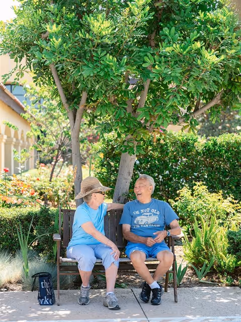 Two elderly people sitting on a wooden bench under a tree in a garden area, smiling and talking. The woman is wearing a wide-brimmed hat, light blue shirt, and light pants, while the man is wearing a blue t-shirt and shorts. The background shows lush greenery and a building with arches.