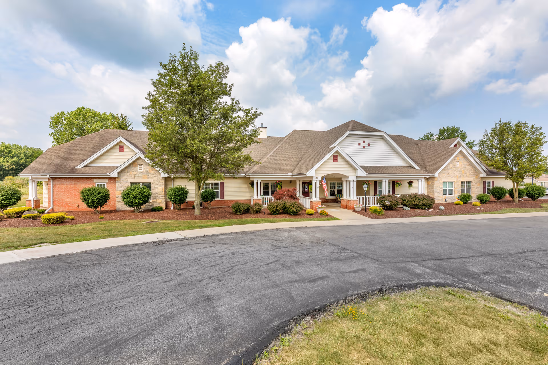 Exterior view of a single-story senior living facility building with a combination of brick, stone, and siding. The building has a covered entrance with white columns, an American flag, and well-maintained landscaping including trees, shrubs, and flower beds. The sky is partly cloudy.