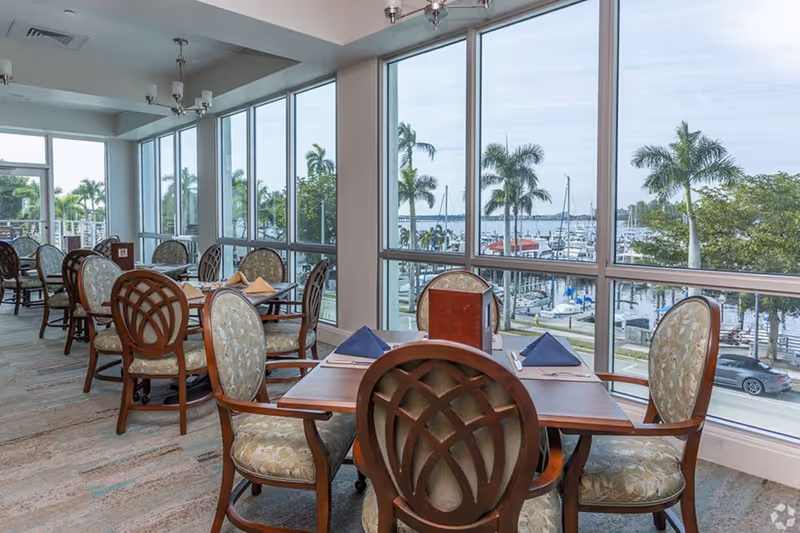 Sunlit dining area with wooden tables and upholstered chairs facing large windows overlooking a marina and palm trees.