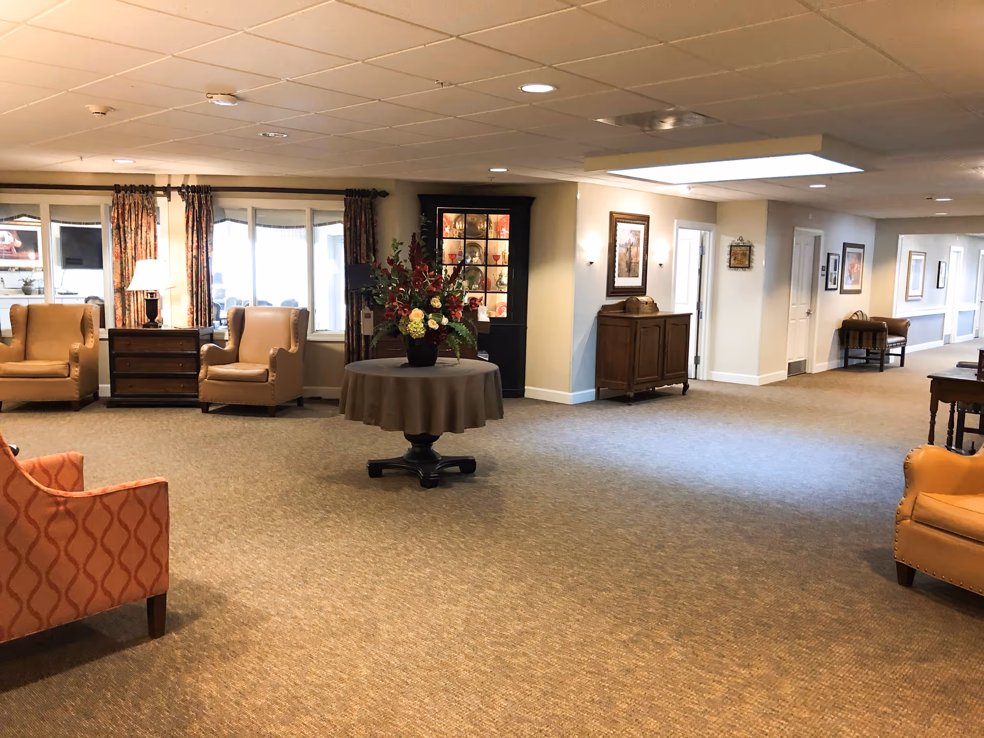 Spacious carpeted common lounge with armchairs, a central round table holding a floral arrangement, cabinets and a corridor leading to rooms.
