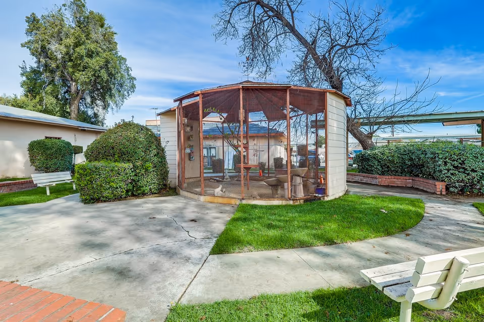 Outdoor garden area at Colonial Gardens Nursing Home featuring a circular screened enclosure with a tree inside, surrounded by green grass, trimmed bushes, a concrete pathway, and white benches. The sky is clear and blue.