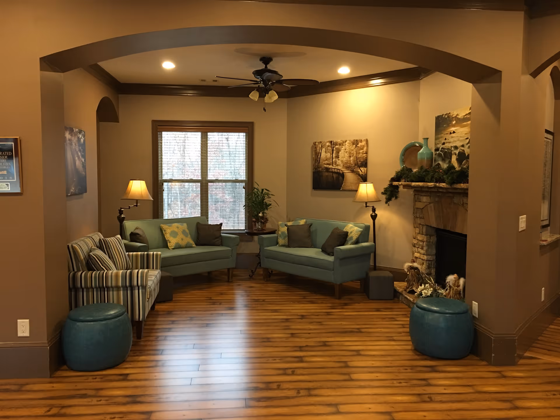 A cozy living room area with wooden flooring, two teal sofas with cushions, a striped armchair, two floor lamps, a ceiling fan with lights, a window with blinds, and a stone fireplace decorated with greenery and vases.
