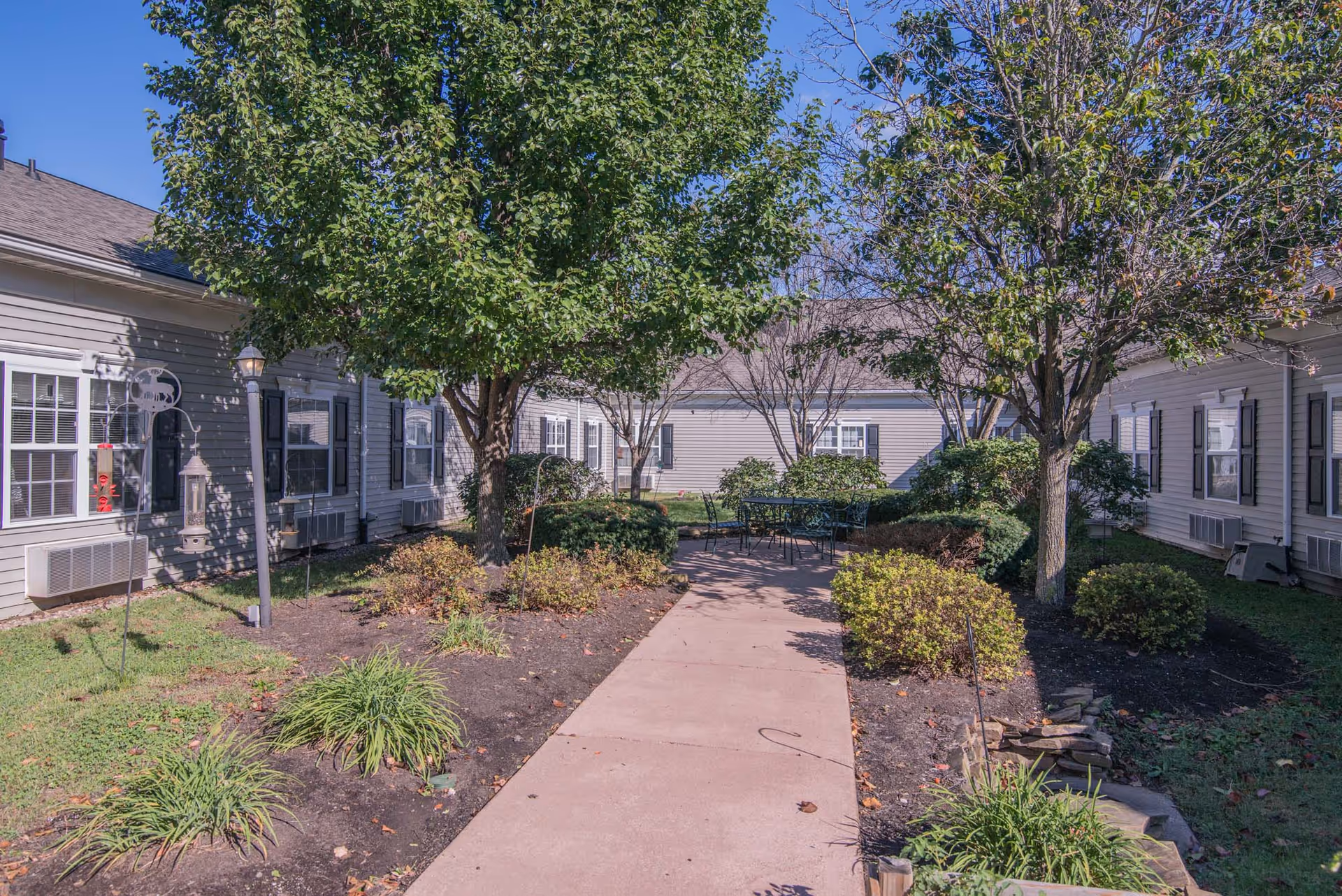 Outdoor courtyard area at Morning Pointe of Russell with a paved walkway, green trees, bushes, and a seating area with metal chairs and tables surrounded by single-story building wings with windows and air conditioning units.