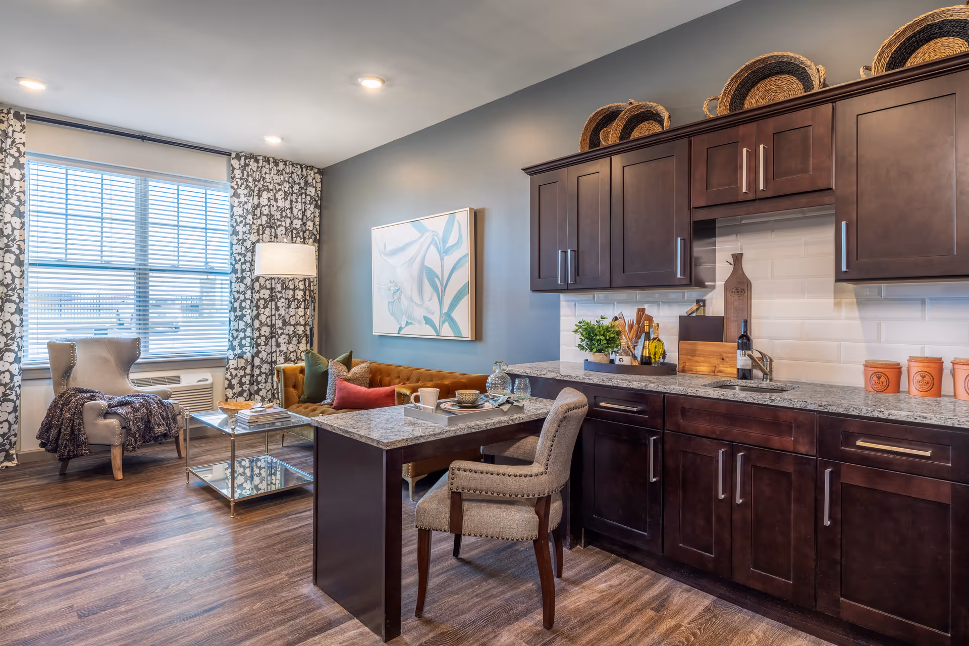 A cozy living area with a small kitchen featuring dark wood cabinets and a granite countertop. The living space includes a brown sofa with colorful pillows, a glass coffee table, an armchair with a throw blanket, and a floor lamp next to a window with patterned curtains. The walls are painted gray, and there is a floral artwork hanging above the sofa.