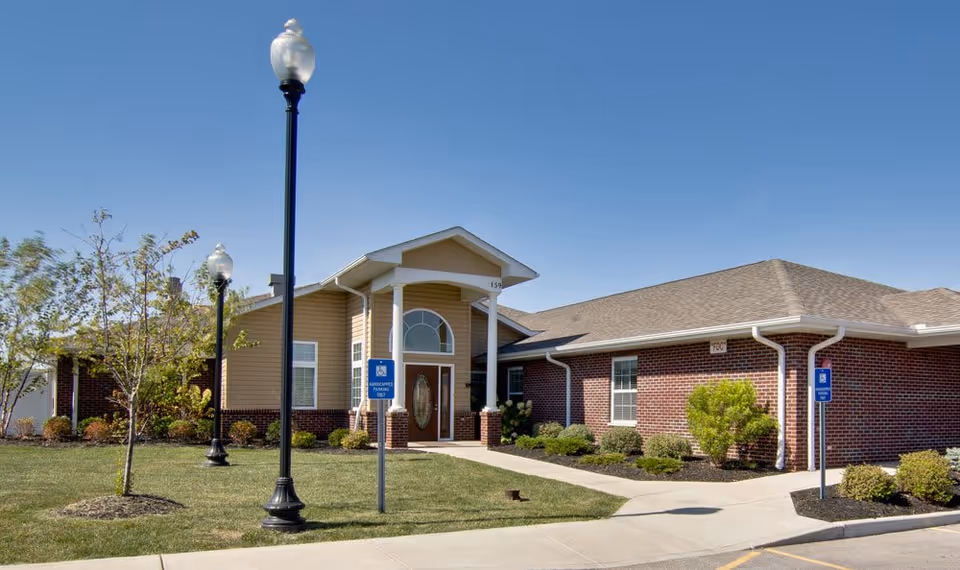 Exterior view of a single-story senior living facility building with a brick and beige siding facade, a covered entrance with white columns, two street lamps, a small tree, and landscaped bushes under a clear blue sky.