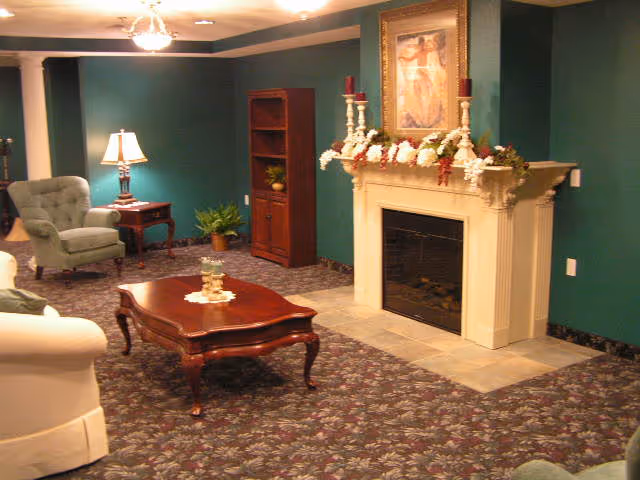 A cozy living room with green walls and floral carpet. The room features a white fireplace decorated with a garland and candles, a wooden coffee table with a doily and candle centerpiece, a green upholstered armchair, a side table with a lamp, a wooden bookshelf, and a framed painting above the fireplace.