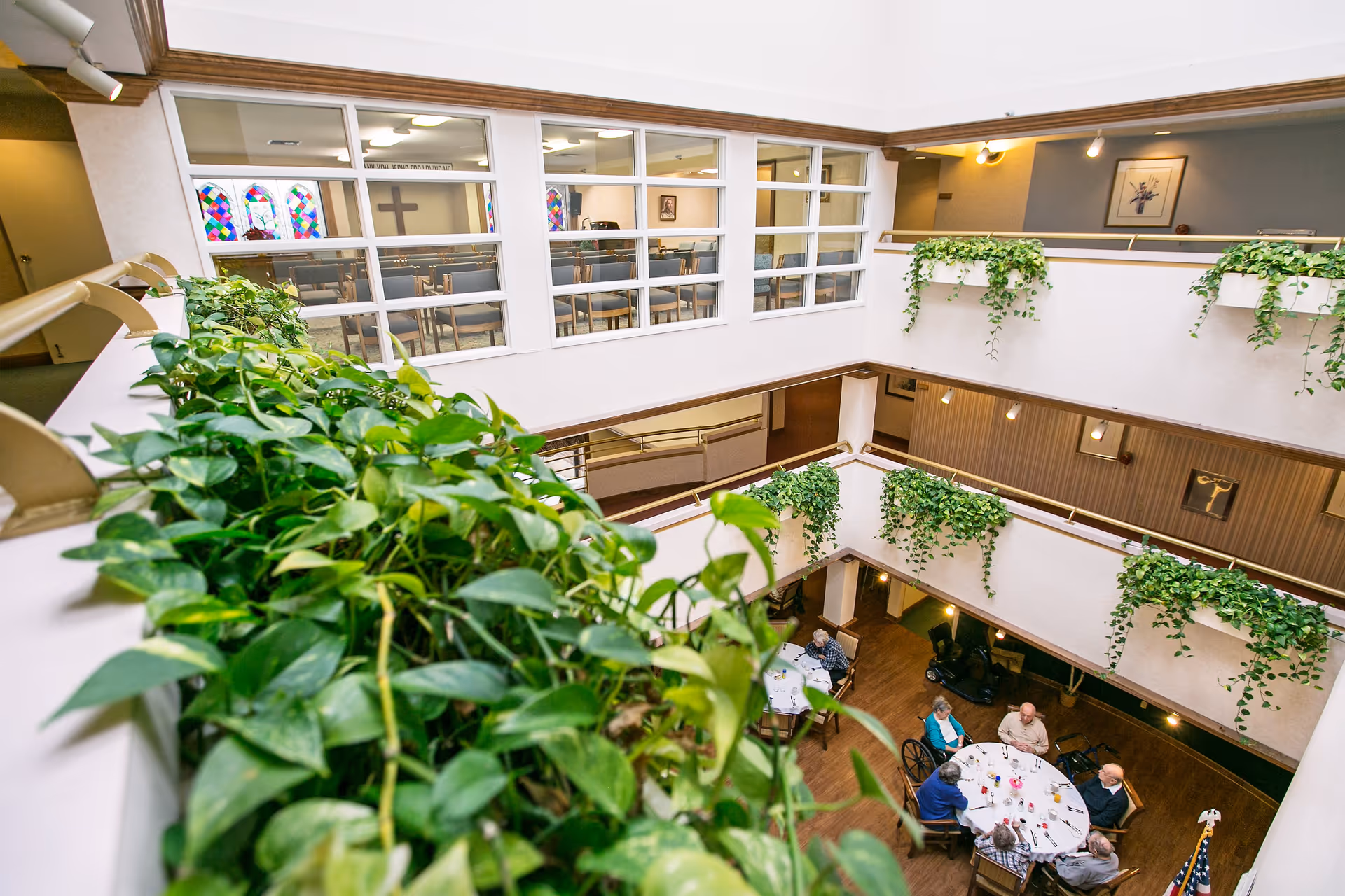 View from an upper floor overlooking a common area in a senior living facility. Several elderly people are seated around a round table engaged in conversation. The area is decorated with green plants hanging over the railings and framed artwork on the walls. Through large windows, a chapel with stained glass windows and a cross is visible in the background.