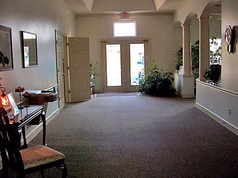 Interior view of a hallway or lobby area with carpeted floor, a chair and small table with decorative items on the left, potted plants near the glass double doors at the far end, and arched openings on the right side wall.