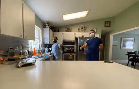 Two masked staff members standing in a bright residential-style kitchen with white cabinets, stainless appliances, and a large countertop in the foreground.