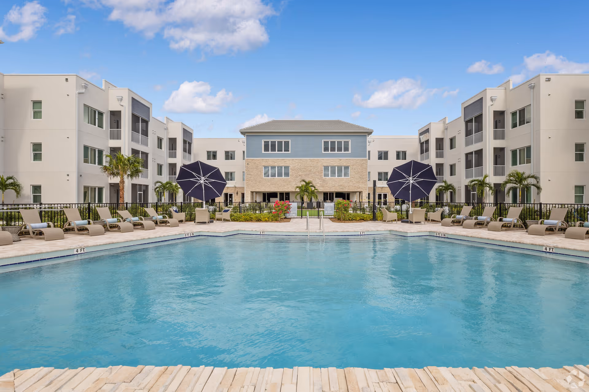 Outdoor swimming pool with lounge chairs and umbrellas surrounding it, set in front of a modern three-story residential building under a blue sky with some clouds.