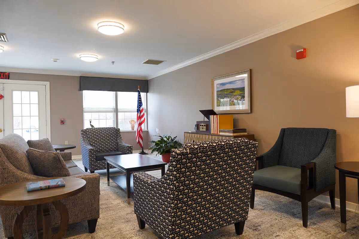 A cozy living room area with a sofa, two patterned armchairs, and a dark upholstered armchair arranged around a rectangular coffee table. There is a side table with a book on it next to the sofa. The room has beige walls, a framed landscape picture, a potted plant on a cabinet, and an American flag near a window with blinds. Ceiling lights illuminate the space.