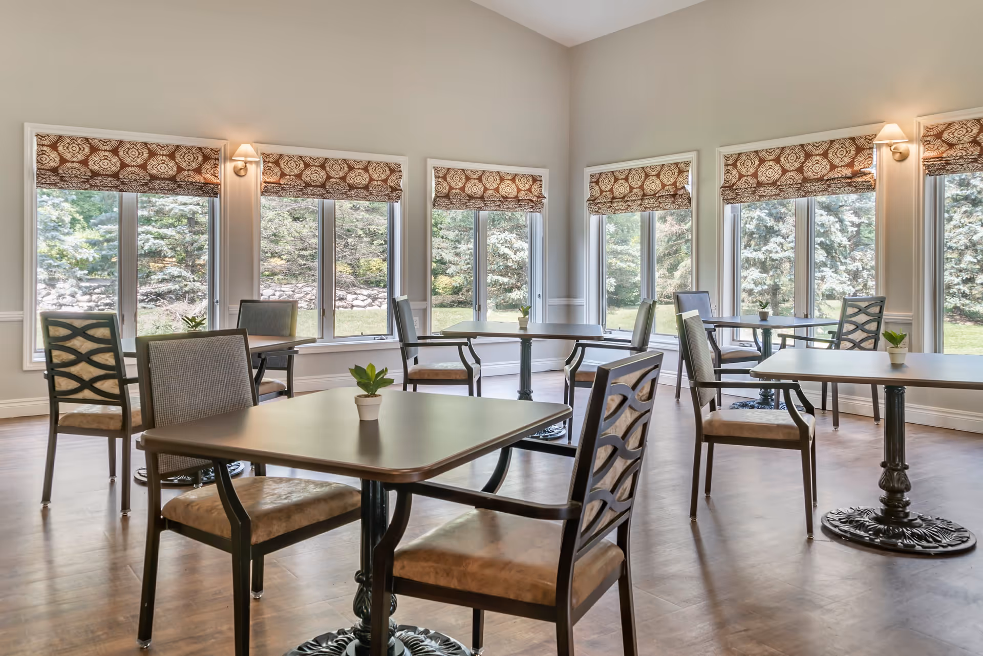 A bright dining room with multiple square tables and chairs arranged neatly. Each table has a small potted plant as decoration. Large windows with patterned Roman shades allow natural light to fill the room and provide a view of greenery outside.