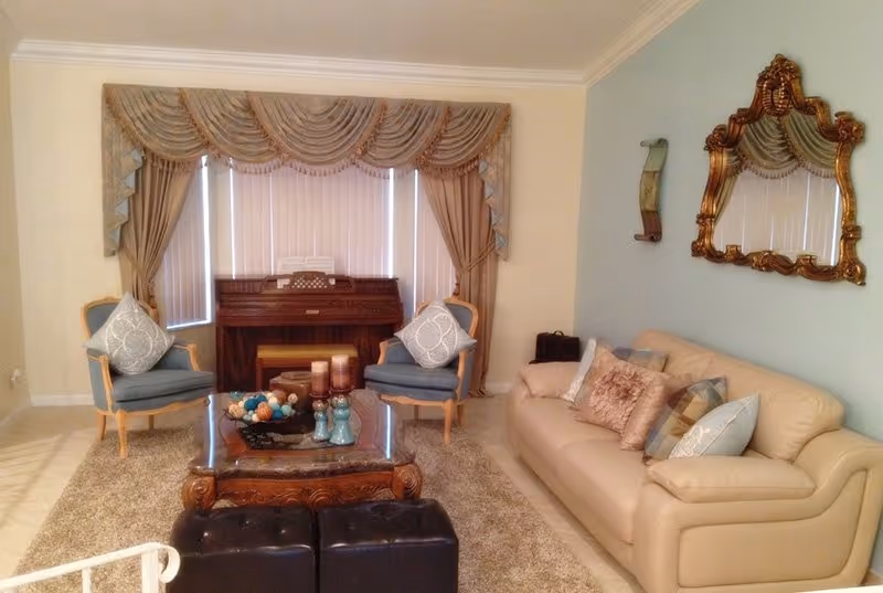 A cozy living room with a beige leather sofa adorned with multiple decorative pillows, two blue cushioned armchairs with patterned pillows, a wooden coffee table with candles and decorative items, a piano against the window with vertical blinds and ornate drapes, a large ornate gold-framed mirror on the wall, and a soft beige carpet covering the floor.