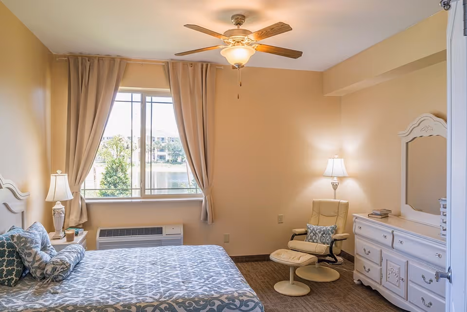 A cozy bedroom with a bed covered in blue and white patterned bedding, a bedside table with a lamp, a window with beige curtains showing an outdoor view, a ceiling fan with a light, a cream-colored armchair with a matching ottoman, and a white dresser with a mirror and a lamp on top.
