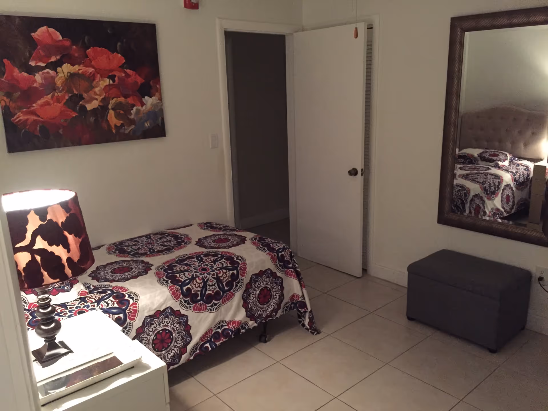 A small bedroom with a single bed covered in a patterned bedspread with purple, pink, and white colors. A bedside table with a lamp featuring a dark patterned shade is next to the bed. A large mirror hangs on the wall above a gray storage ottoman. A floral painting with red and pink flowers is mounted on the wall above the bed. The room has tiled flooring and a door that is partially open.