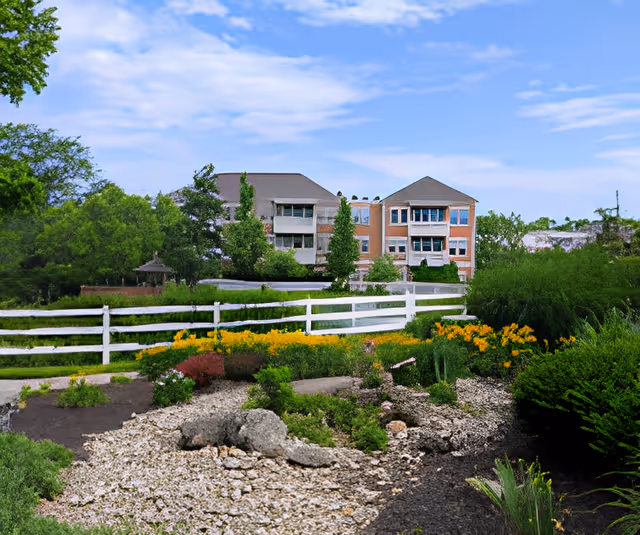 Brick multi-story senior living building viewed across a landscaped garden with a white split-rail fence and flowering plants under a blue sky.