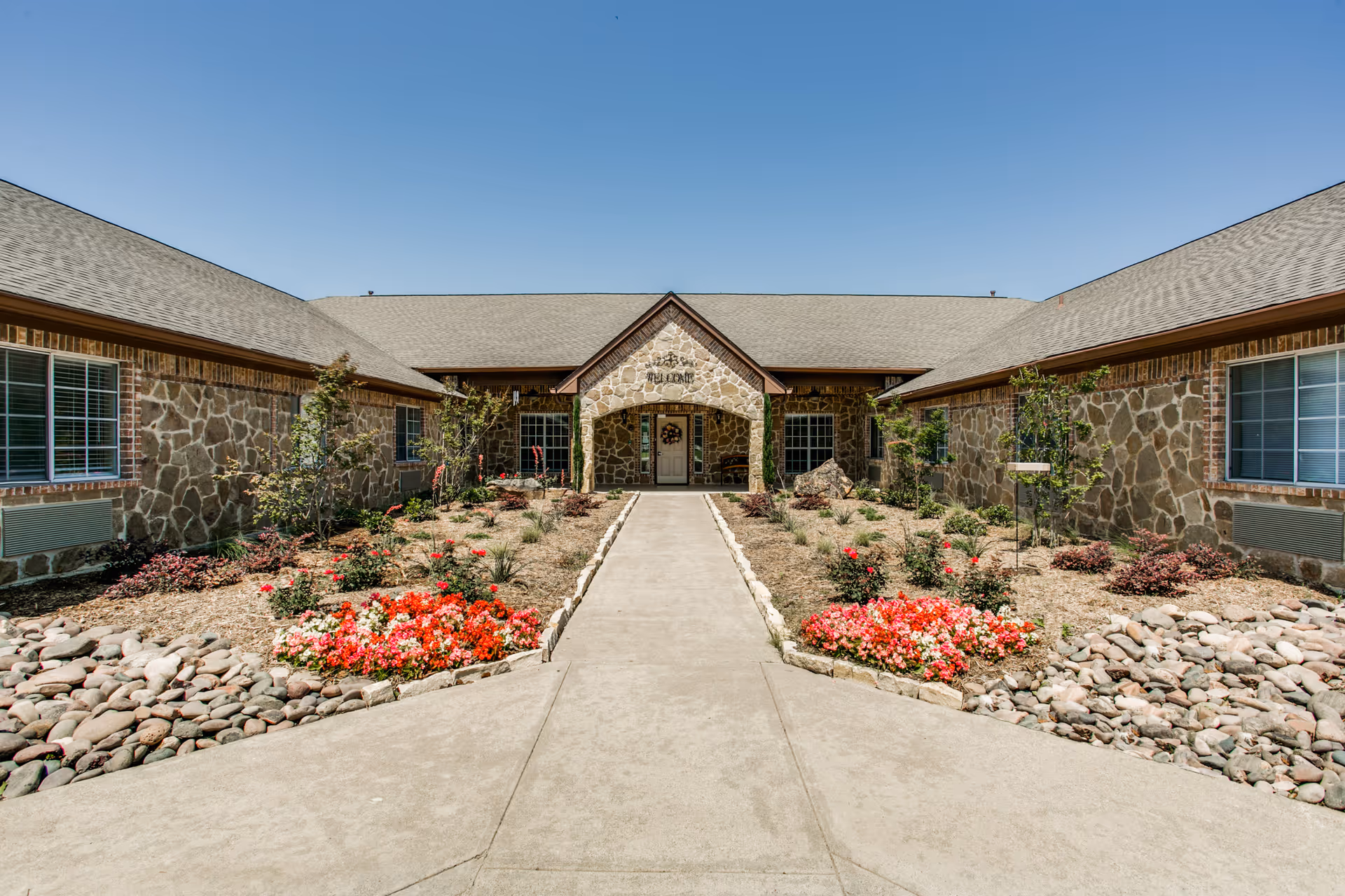 Front exterior view of Avalon Memory Care facility with a stone facade, a central entrance with a covered porch, landscaped flower beds with red and pink flowers, and a clear blue sky overhead.