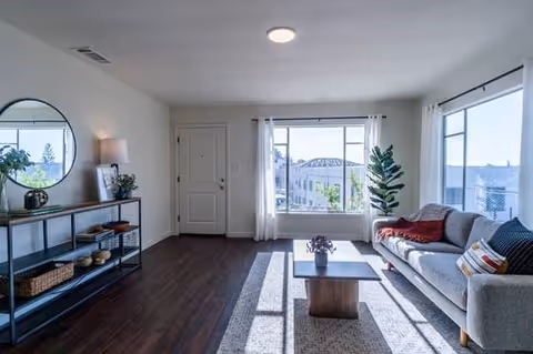 A bright living room with large windows letting in natural light. The room features a gray sofa with colorful pillows and a red throw blanket, a wooden coffee table with a small plant, a patterned rug, and a console table with decorative items including a round mirror, lamp, and plants. The floor is dark wood, and the walls are painted white.