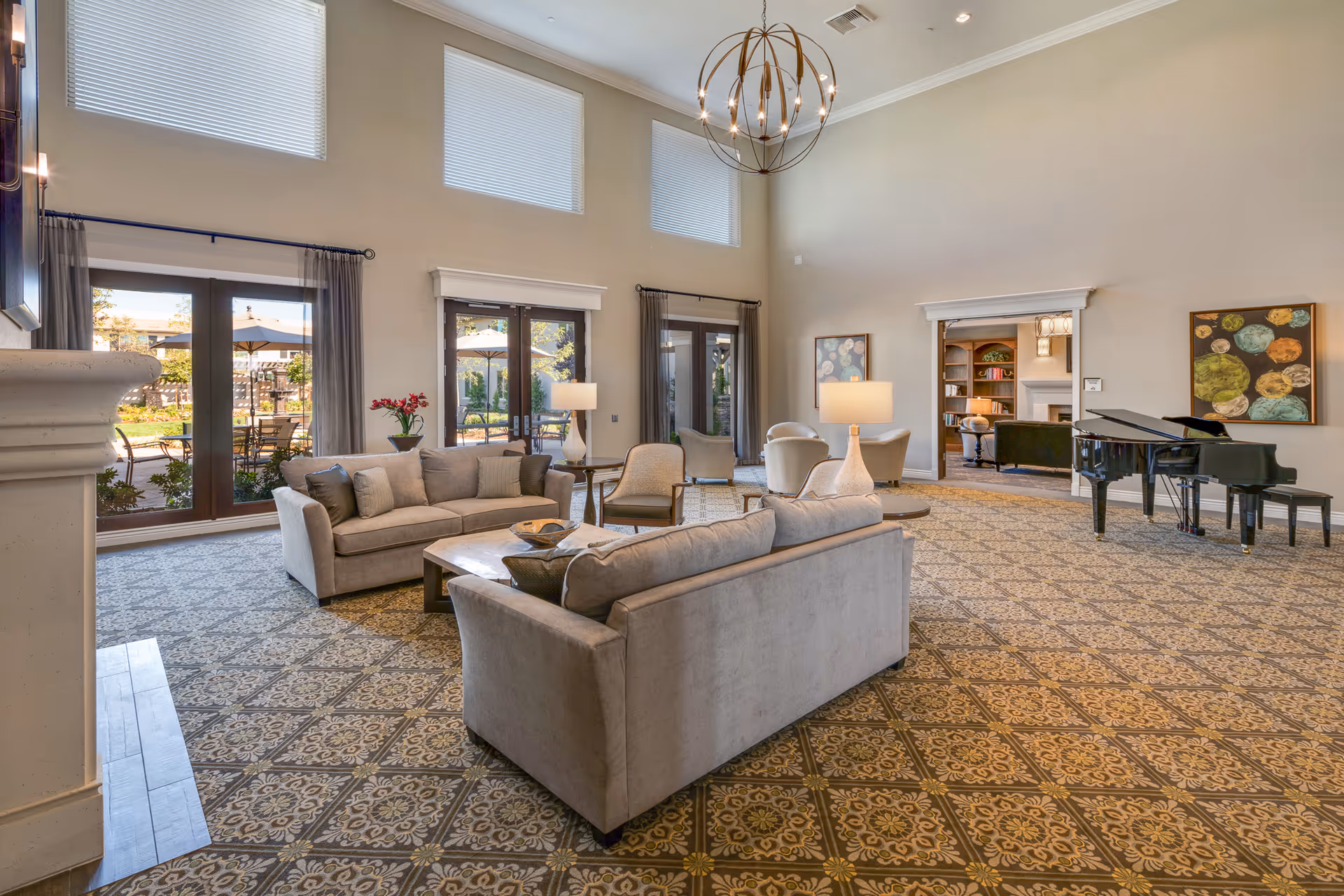 Spacious, well-lit common lounge with sofas, armchairs, a coffee table, grand piano, tall windows, and views to an outdoor patio.