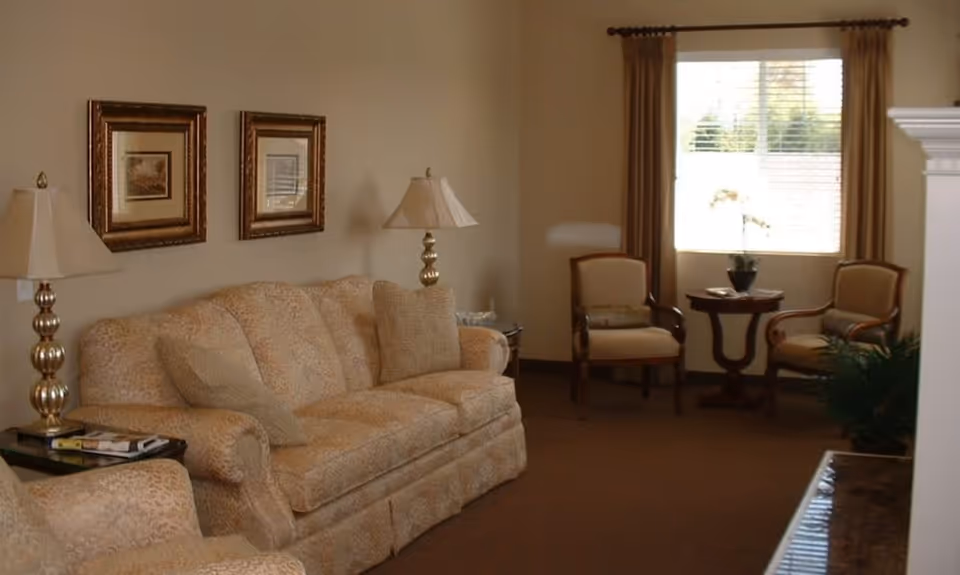 A cozy living room with a beige patterned sofa and matching armchair, two table lamps with gold bases, two framed pictures on the wall, two wooden armchairs with beige cushions, a small round wooden table with a potted plant, and a window with brown curtains letting in natural light.
