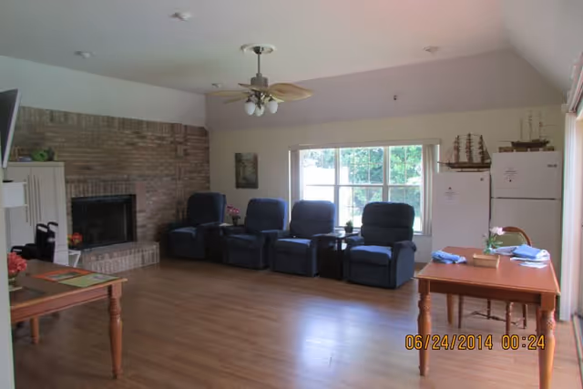 A living room area with four blue recliner chairs lined up against a wall with a large window. There is a brick fireplace on the left side and two wooden tables with chairs on the right side. A ceiling fan with lights is mounted on the ceiling. The floor is wooden, and there are decorative items including model ships on top of two white refrigerators near the window.