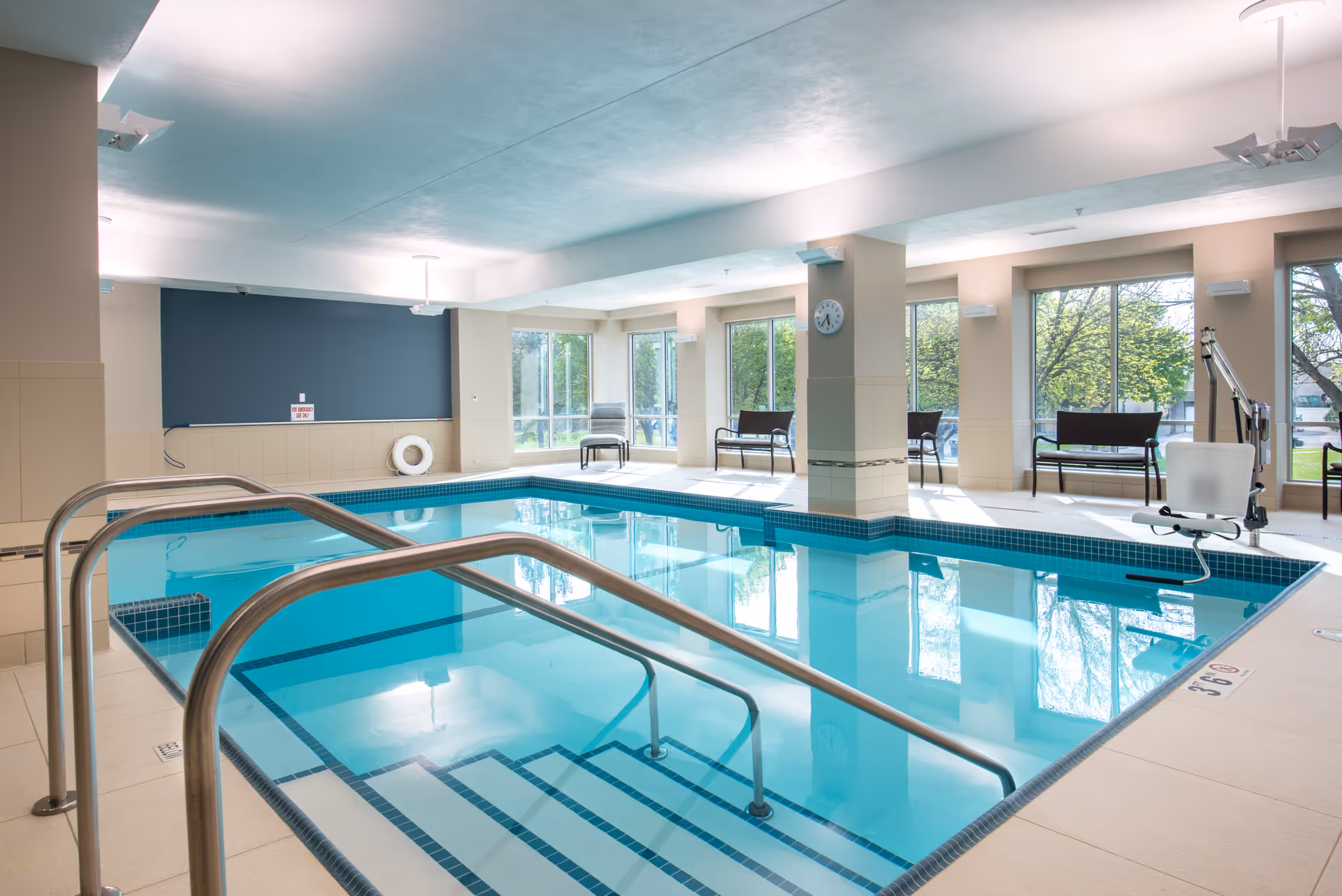 Indoor swimming pool area with handrails leading into the water, large windows letting in natural light, benches along the walls, and a pool lift for accessibility.