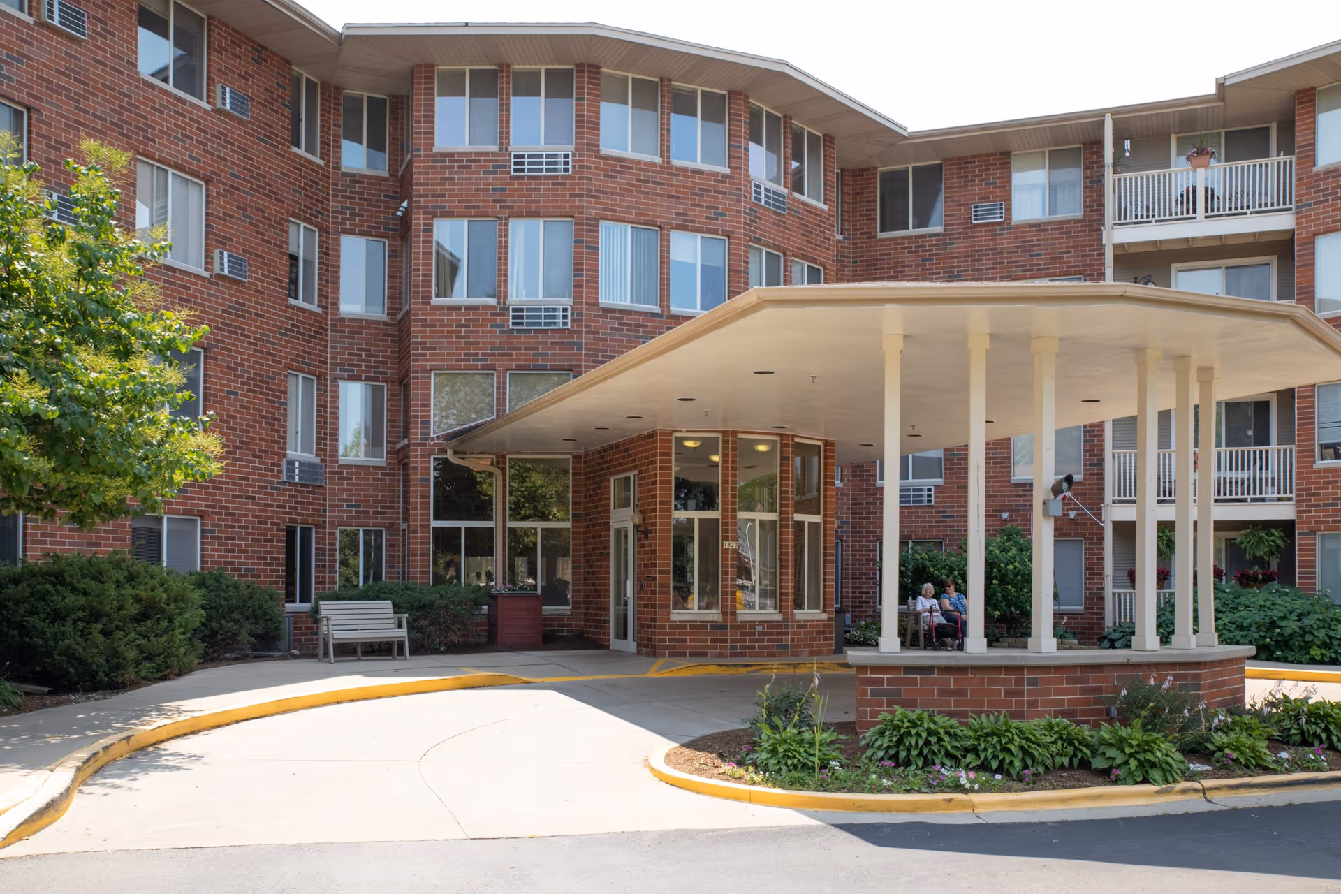 Exterior view of a multi-story brick senior living facility with a covered entrance supported by white columns. There are windows and balconies visible, along with some greenery and a bench near the entrance. Two elderly people are sitting under the covered area.