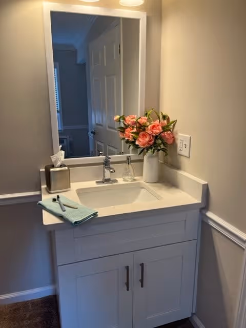A small bathroom vanity with a white countertop and sink. On the countertop, there is a vase with pink flowers, a soap dispenser, a tissue box, and a folded green towel with a toothbrush on top. A large mirror is mounted on the wall above the sink, reflecting a partially open door and part of another room.