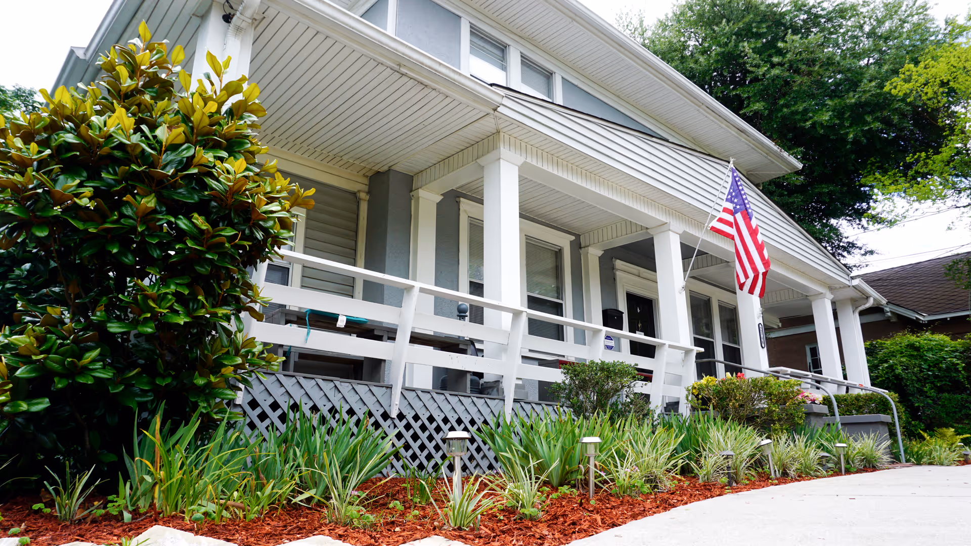 Exterior view of a single-story house with a white porch railing, an American flag mounted near the entrance, and well-maintained landscaping with green shrubs and plants along a concrete walkway.