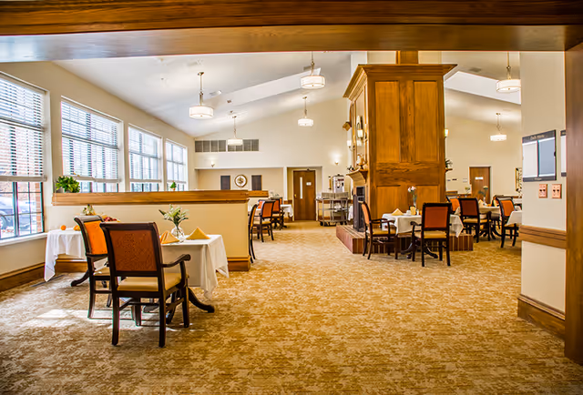 Spacious assisted-living dining room with multiple set tables and chairs, large windows, and warm wood accents.
