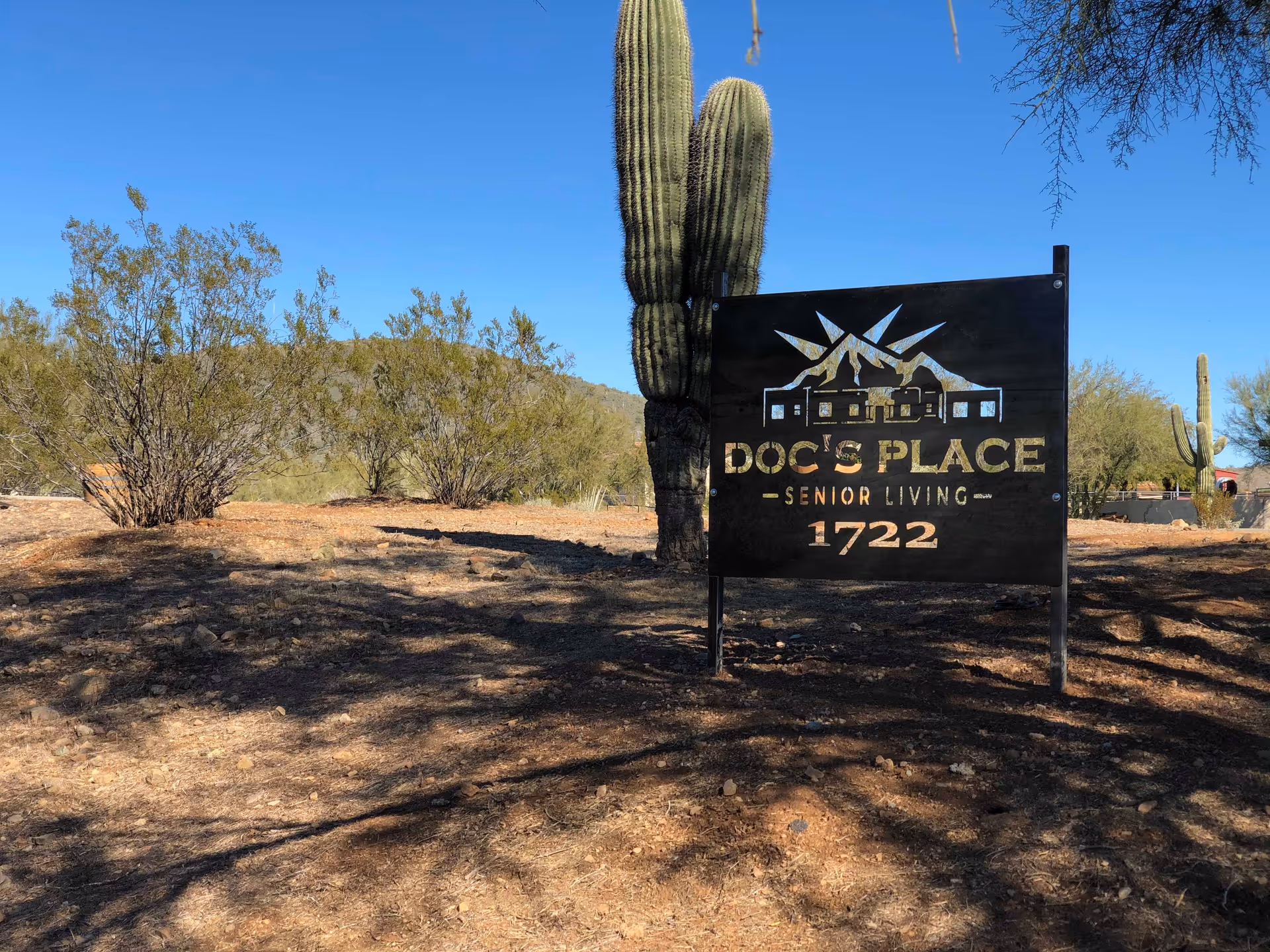 A metal sign reading "Doc's Place Senior Living 1722" stands in a desert landscape with saguaro cacti and a clear blue sky.