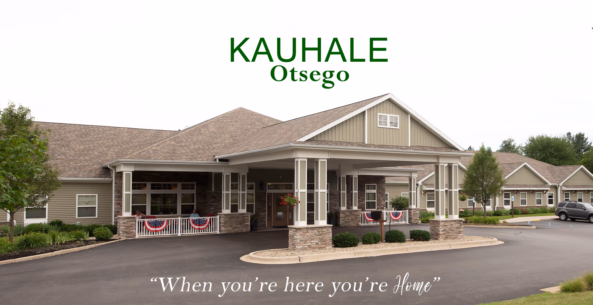 Exterior view of Kauhale Otsego senior living facility showing a single-story building with a covered entrance, stone pillars, and American flag decorations on the railings. There are trees and shrubs around the building and a parking lot in front.
